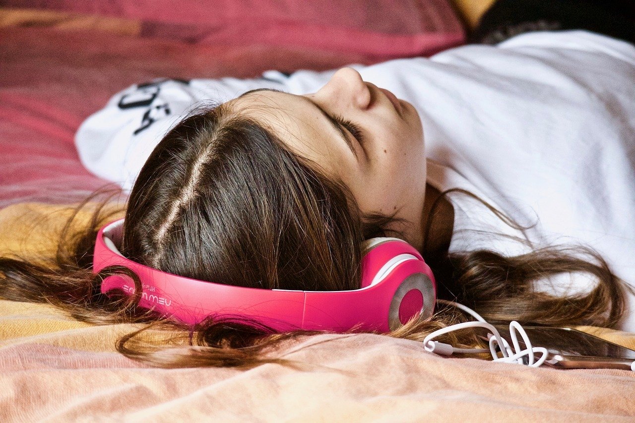 Ragazza stesa sul letto ad ascoltare podcast con delle cuffie rosa