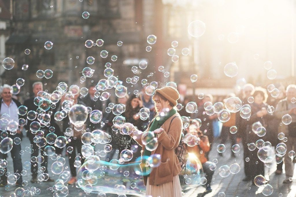 Fotografia di una giovane donna che gioca con bolle di sapone in una piazza affollata, illuminata dalla luce del sole. L’immagine trasmette leggerezza, spensieratezza e gioia condivisa in un contesto urbano.