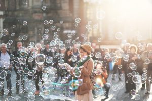 Fotografia di una giovane donna che gioca con bolle di sapone in una piazza affollata, illuminata dalla luce del sole. L’immagine trasmette leggerezza, spensieratezza e gioia condivisa in un contesto urbano.