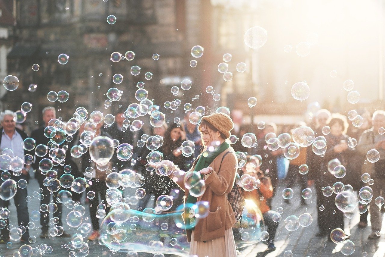 Fotografia di una giovane donna che gioca con bolle di sapone in una piazza affollata, illuminata dalla luce del sole. L’immagine trasmette leggerezza, spensieratezza e gioia condivisa in un contesto urbano.