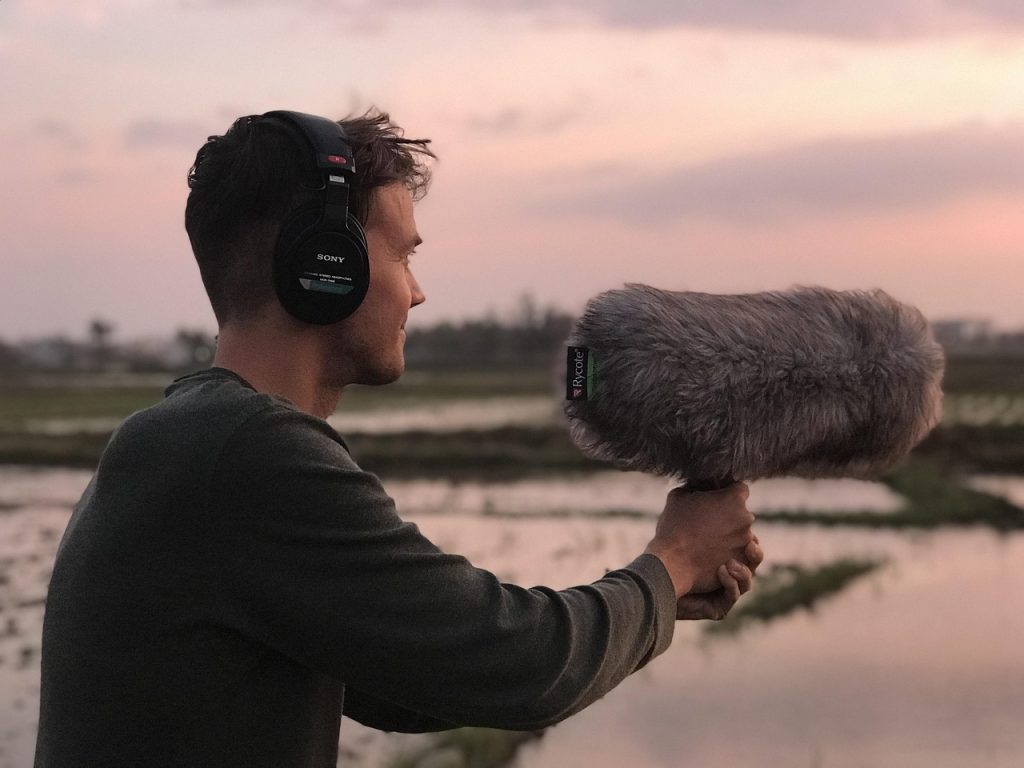 Un tecnico del suono con cuffie Sony registra l’audio all’aperto utilizzando un microfono direzionale con antivento in una zona naturale al tramonto. L’immagine rappresenta il lavoro sul campo di chi produce podcast, documentari e registrazioni ambientali.